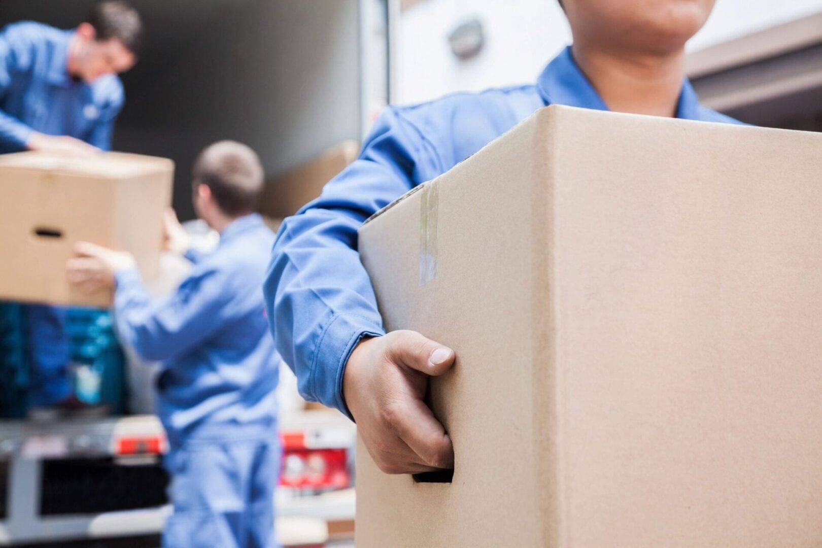 Three Young Delivery Men Carrying Cardboard Box In Front Of Truck